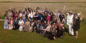 A large group of delegates at the Arts Health Research Intensive are outside in a grassy landscape with blue sky, many are waving and smiling.
