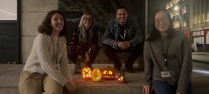 Four people are gathered around a set of carved, illuminated pumpkins displayed on a ledge in an outdoor, urban setting at night. The group is smiling and the surrounding buildings are dimly lit in the background, adding to the festive atmosphere.