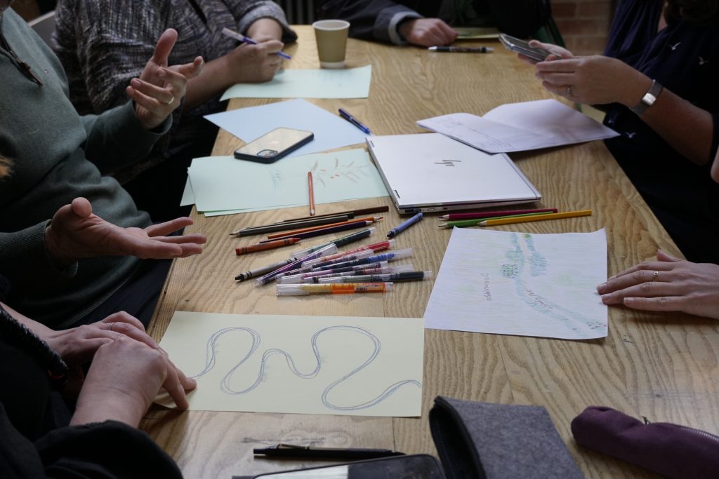 A photo shows a table with paper, pens and drawings on. People sit around its edges and we see their hands in motion. 