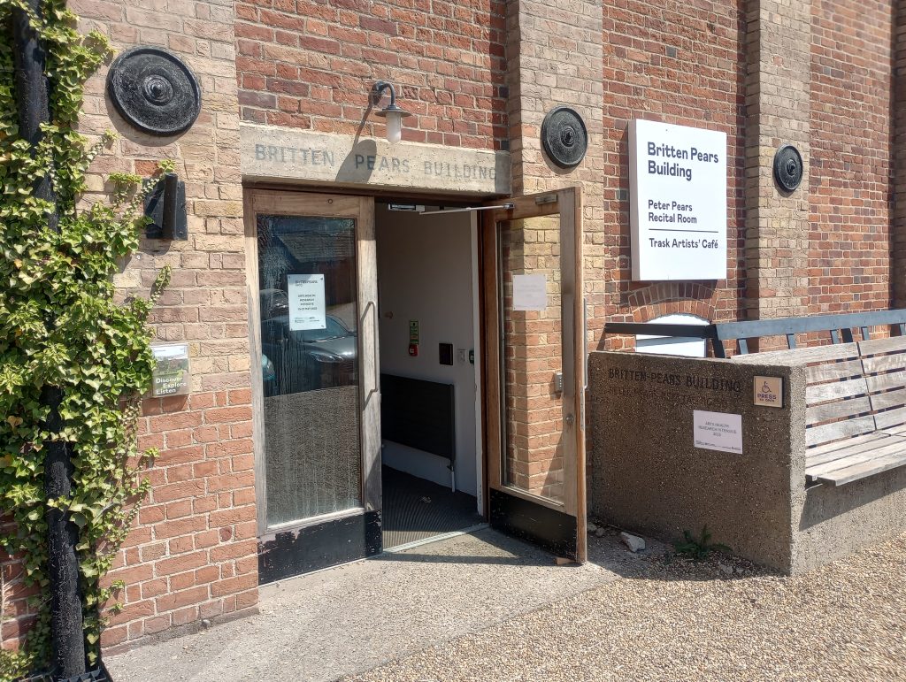 A sunny photo shows a brick wall with an open doorway. A sign indicates that it is the "Britten Pears Building". 