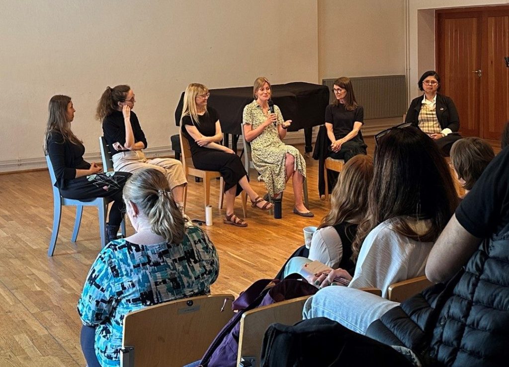 Members of the Arts Health Research Intensive faculty sit at the front of a lecture theatre. One person speaks with a microphone whilst others listen. 